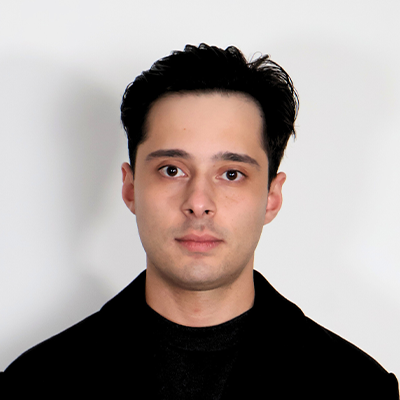 Headshot of chiropractor Mustafa Shalal with dark hair, wearing a black coat against a light gray background.